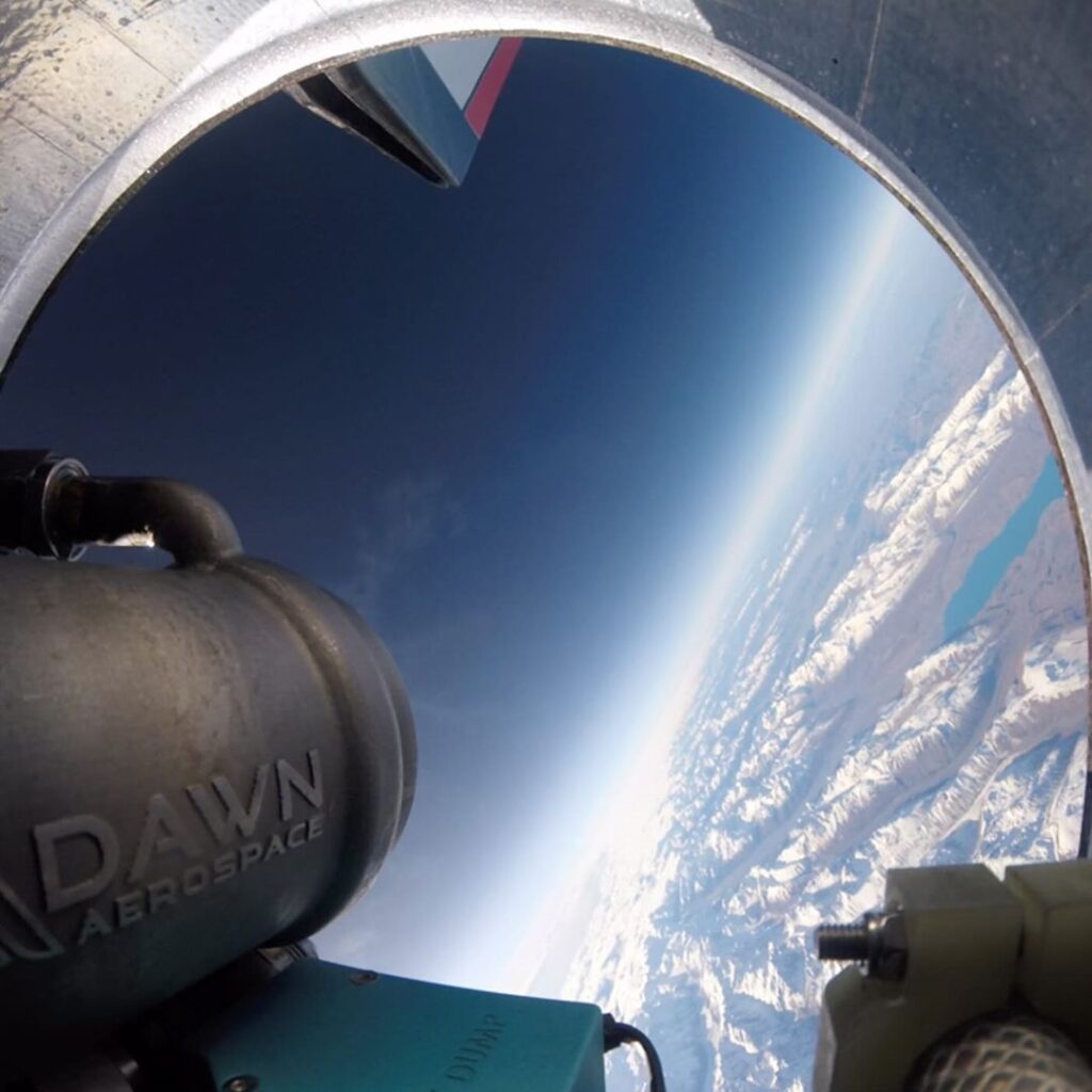 O Mk-II Aurora no céu, com a câmera capturando a saída do motor e o logo da Dawn Aerospace visível. Ao fundo, as montanhas parcialmente cobertas de gelo formam uma paisagem impressionante, destacando o voo supersônico do jato