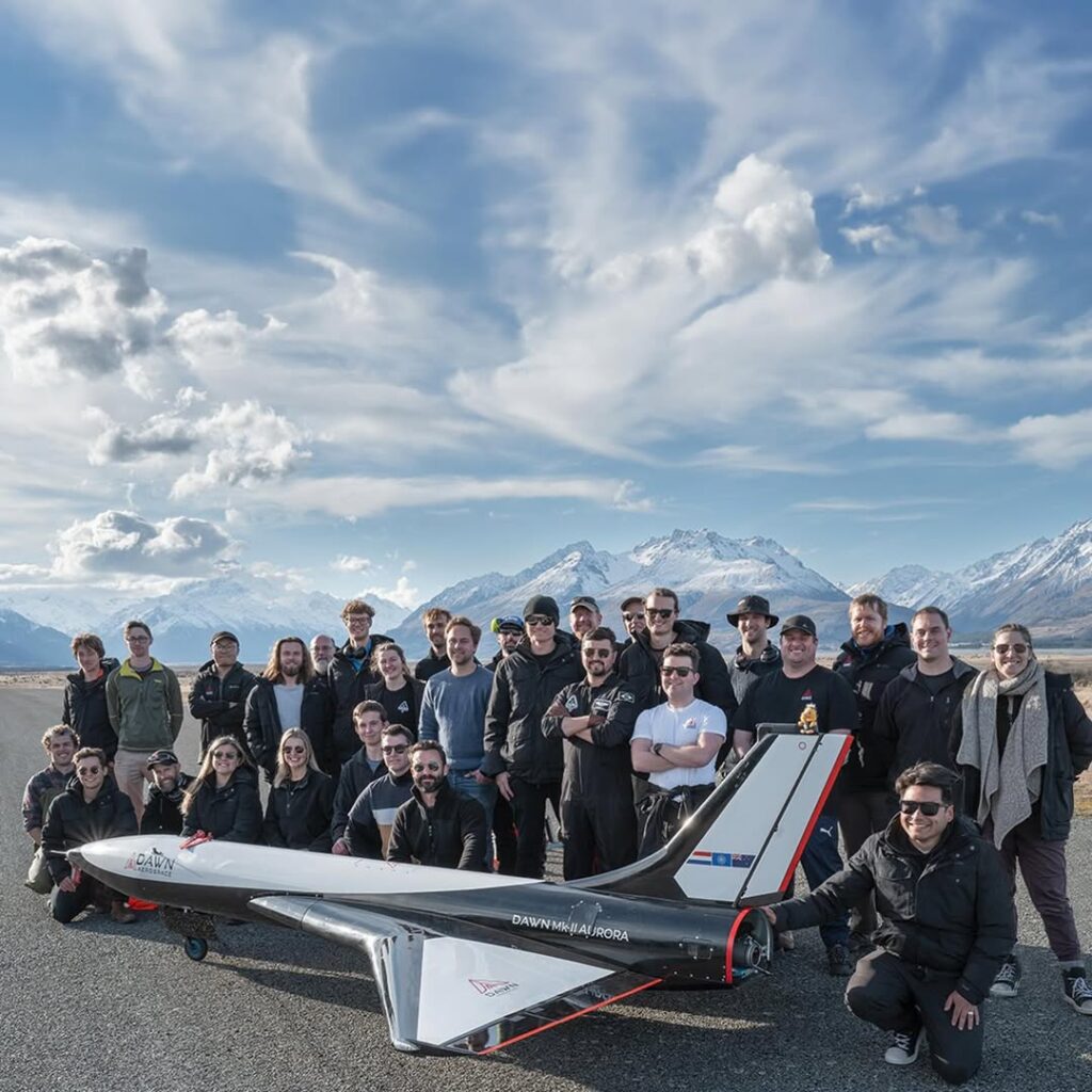 Equipe de cerca de 30 pessoas posando para uma foto com o Mk-II Aurora ao fundo. O jato, com a bandeira da Nova Zelândia e o logo da empresa Dawn Aerospace, está estacionado em uma pista com as montanhas da Nova Zelândia visíveis ao fundo durante o dia, destacando a conquista histórica do voo supersônico civil."