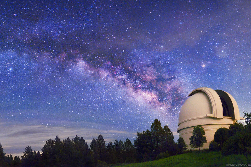 O famoso telescópio do Monte Palomar, com sua icônica cúpula branca destacando-se contra o céu estrelado. Ao fundo, é visível uma galáxia distante, refletindo a importância histórica e científica deste observatório.