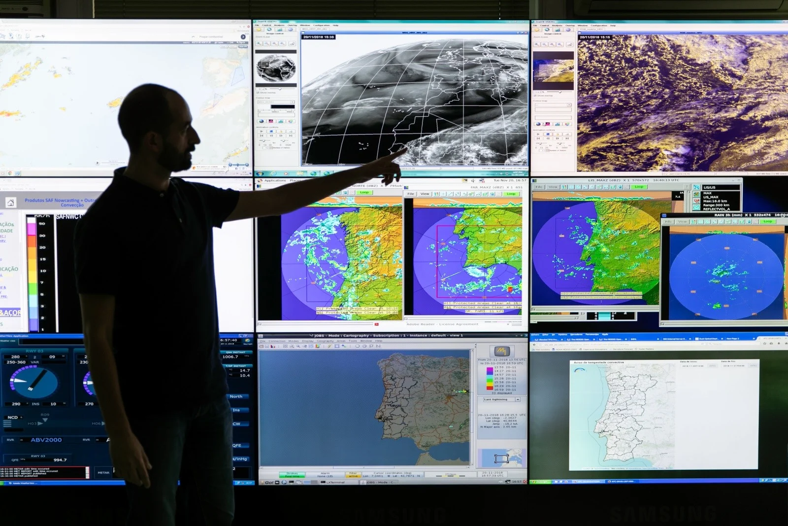 Homem meteorologista em frente a vários monitores exibindo previsões de clima e do tempo, representando o trabalho de monitoramento e análise meteorológica.