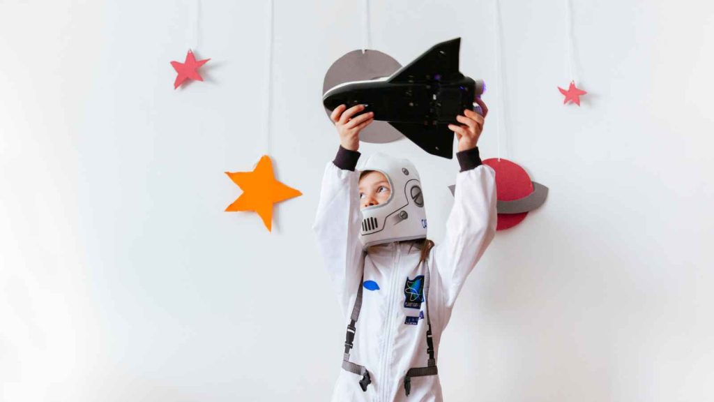 Criança com traje de astronauta branco brincando com um foguete de brinquedo, com recortes de planetas e estrelas na parede branca ao fundo.