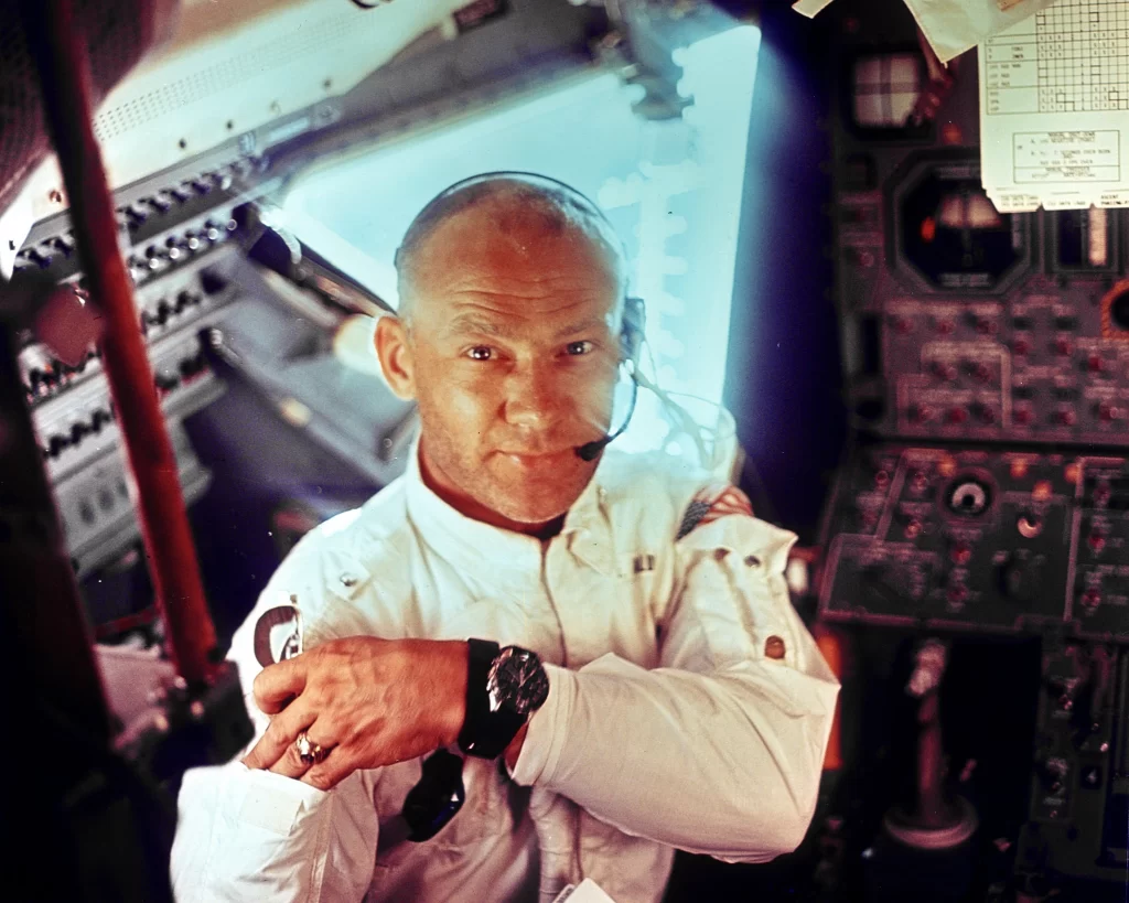 Buzz Aldrin a bordo do módulo lunar Eagle durante a missão Apollo 11. Ele está de frente para a câmera, usando traje espacial branco e um relógio preto, sem capacete. Ele parece estar guardando algo no bolso de seu traje, em um momento único a bordo da nave lunar.