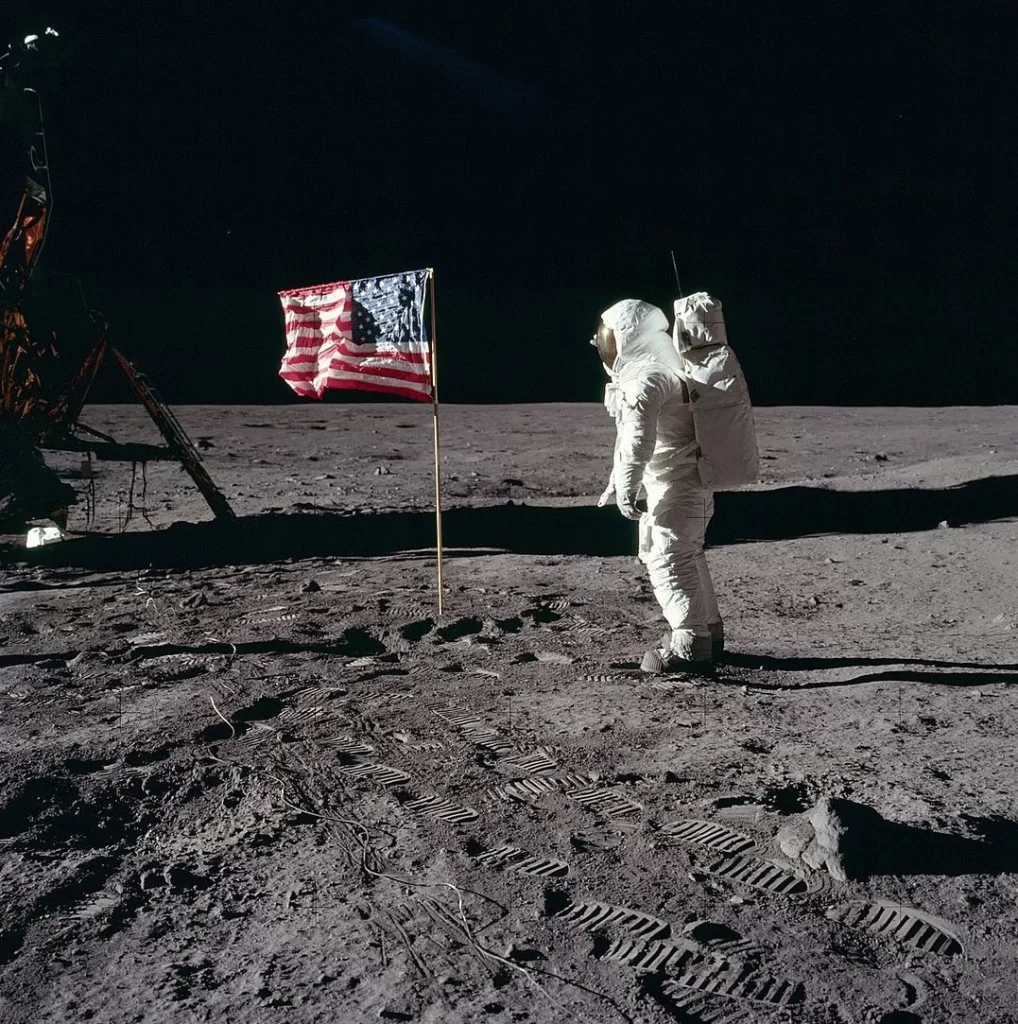 Buzz Aldrin saúda a bandeira americana na Lua durante a missão Apollo 11, em 1969. Ele está de traje espacial completo, com a bandeira americana plantada no solo lunar ao lado dele, simbolizando o marco histórico do pouso lunar dos Estados Unidos.