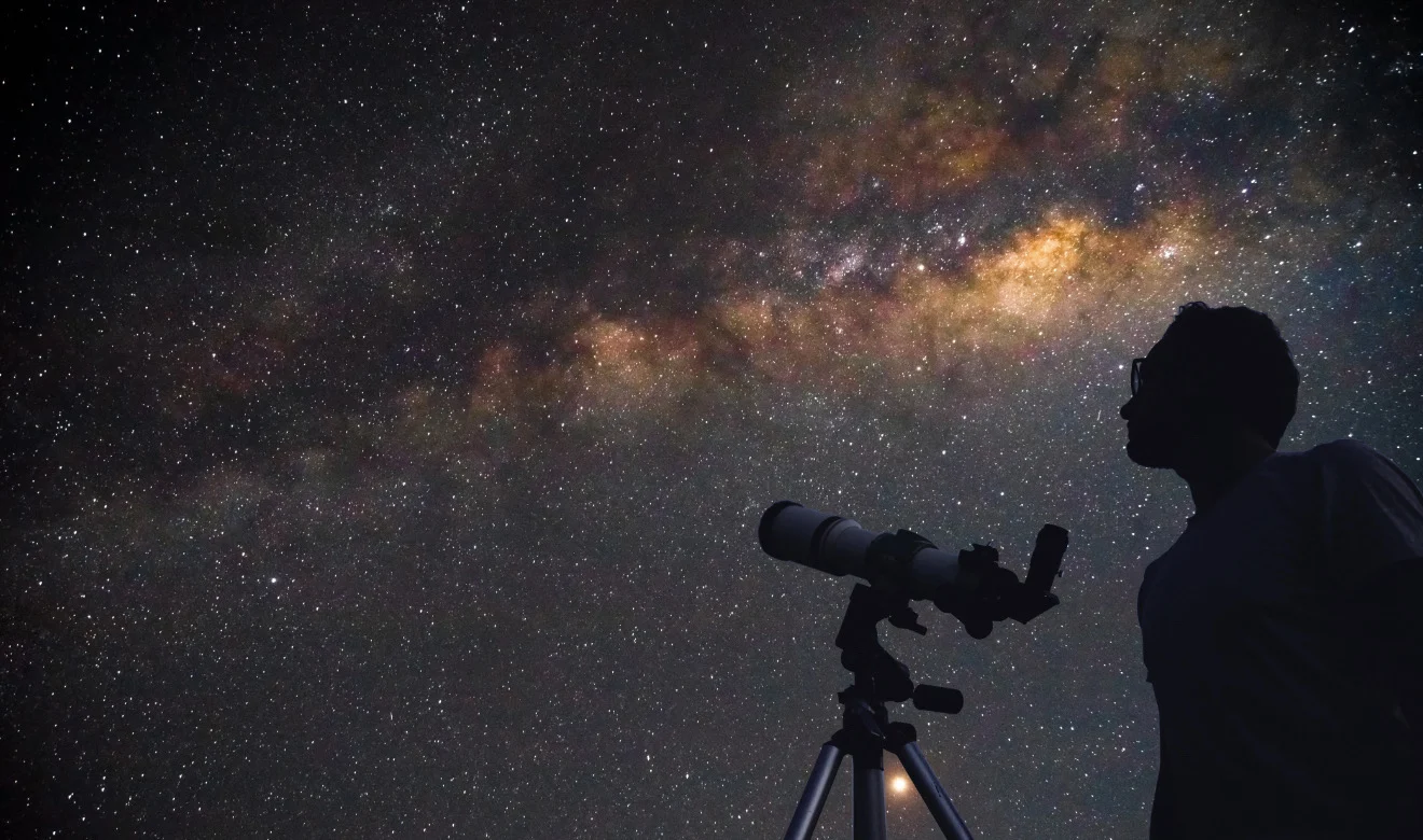Astrônomo com telescópio apontado para o céu noturno estrelado. A imagem ilustra como observar o céu e apreciar fenômenos como chuvas de meteoros e estrelas cadentes.