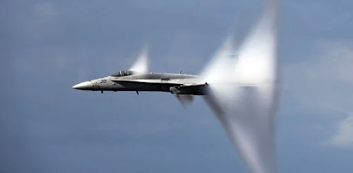 caça de combate quebrando a barreira do som, formando um anel de vapor ao seu redor devido ao efeito sônico. A aeronave atravessa o céu em alta velocidade, deixando um rastro dinâmico, representando o conceito de velocidade supersônica