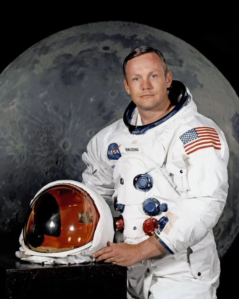 Retrato oficial da NASA de Neil Armstrong, o primeiro homem a pisar na Lua. Ele veste um traje espacial branco, sem capacete, com as mãos apoiadas sobre uma mesa onde seu capacete repousa. Ao fundo, uma imagem da Lua complementa a cena.