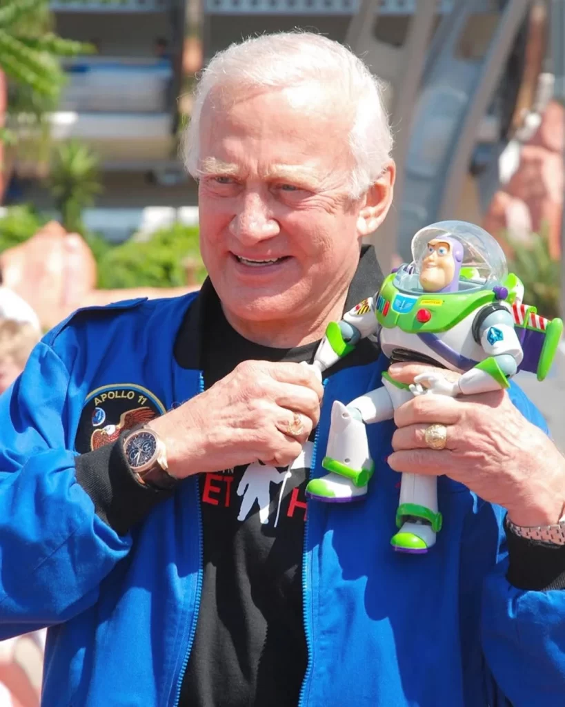 Buzz Aldrin, já idoso, sorrindo enquanto segura um boneco do Buzz Lightyear. Ele está vestindo um traje azul da NASA, com o logo da missão Apollo 11 visível. O boneco de Buzz Lightyear está com traje espacial branco e detalhes em verde e roxo, com o logo da Star Command no peito e as asas nas costas. Esse momento captura um lado descontraído do astronauta, em contraste com sua icônica missão lunar.