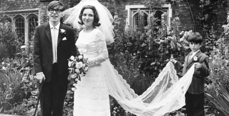 Stephen Hawking e Jane Wilde em seu casamento, em 1965. Stephen está vestindo um terno, enquanto Jane usa um vestido longo. Eles estão sorrindo juntos, capturando um momento de felicidade.