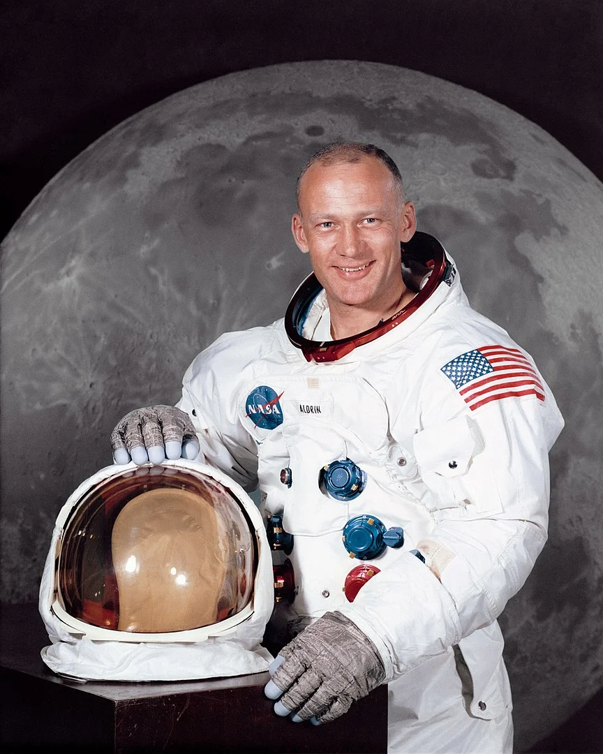 Foto oficial da NASA de Buzz Aldrin, o segundo homem a pisar na Lua durante a missão Apollo 11, sorrindo enquanto está de traje espacial branco, sem capacete. A imagem captura a Lua ao fundo, com suas mãos apoiadas no capacete, marcando um dos momentos mais icônicos da história da exploração espacial.