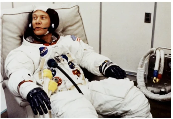Buzz Aldrin de traje espacial branco, sentado em um sofá sem capacete, olhando para a esquerda. A imagem mostra um momento descontraído e tranquilo antes de sua missão histórica, capturando a essência de sua preparação para a exploração espacial.