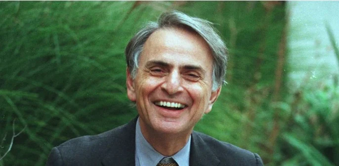 Fotografia de Carl Sagan já adulto, com cabelos grisalhos, vestindo um terno azul escuro e gravata. Ele exibe um sorriso cativante, transmitindo sua paixão pela ciência e pela comunicação do conhecimento astronômico.