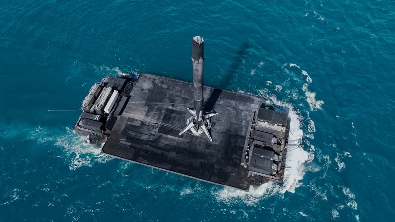 O primeiro estágio do Falcon 9 da SpaceX pousado com sucesso na balsa-drone em meio ao oceano azul-esverdeado. A estrutura metálica do booster exibe marcas de fuligem do intenso calor gerado durante a reentrada. A plataforma flutuante, equipada com sistemas de estabilização, mantém a posição no mar, garantindo a recuperação segura do foguete para reutilização em futuras missões. Ao fundo, o horizonte do oceano se mistura ao céu claro, destacando o feito da engenharia aeroespacial moderna.