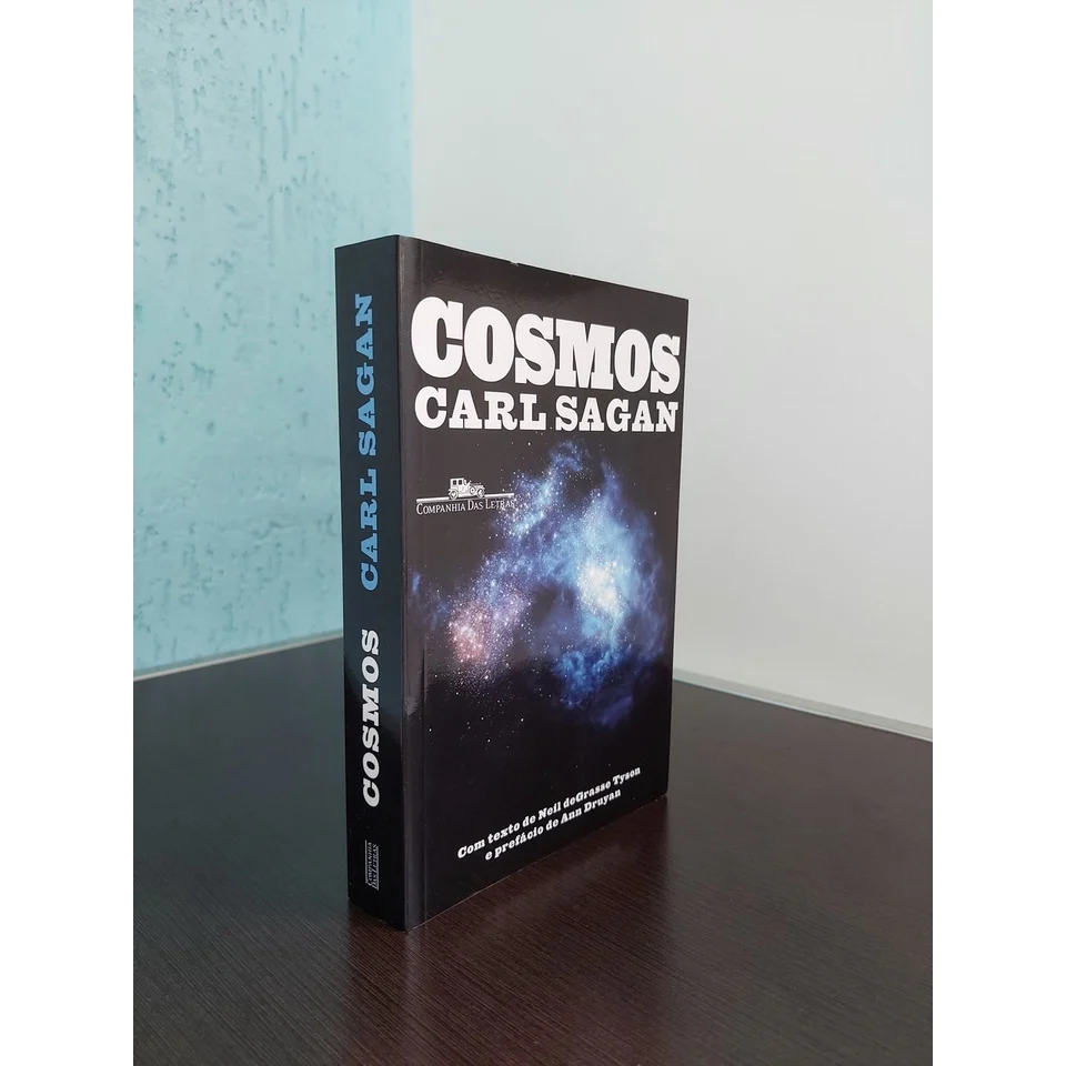 Livro Cosmos, de Carl Sagan, sobre uma mesa de tampo de madeira escura. A capa preta do livro destaca o título em letras brancas, assim como o nome do autor. Ao fundo, uma parede azul com textura adiciona profundidade à cena.