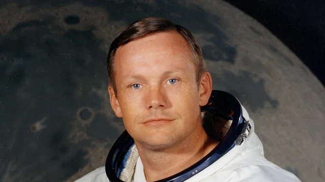 Foto de perfil de Neil Armstrong com traje espacial sem o capacete, em um fundo branco que parece uma sala. A bandeira americana pode ser vista na extremidade do ombro esquerdo do traje.