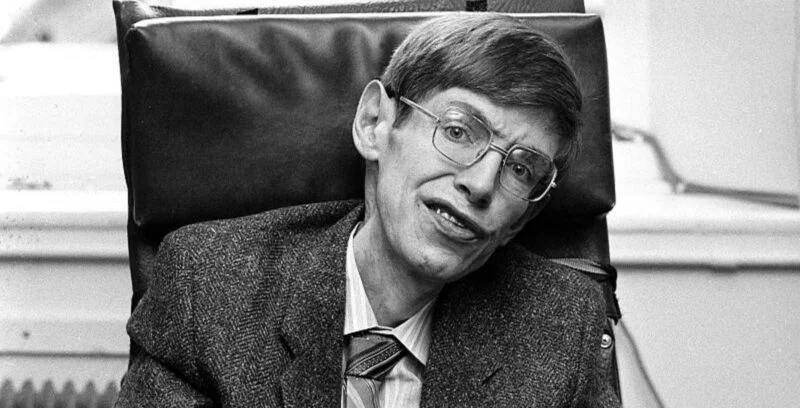 Fotografia em preto e branco de um jovem Stephen Hawking, usando óculos de armação grossa e um paletó. Seu olhar atento e expressão séria refletem sua inteligência e dedicação à física teórica. A imagem captura um momento de sua juventude, antes do avanço dos sintomas de sua condição neuromotora.