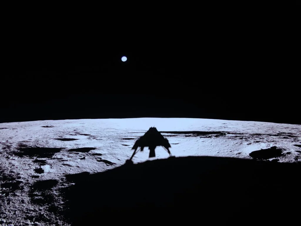 Imagem impressionante mostrando a sombra do lander B Ghost projetada na superfície lunar, com a Terra visível no horizonte. Esta captura marcante simboliza um momento crucial da missão, destacando a relação entre o módulo de pouso e o ambiente lunar, com a majestosa visão da Terra ao fundo.