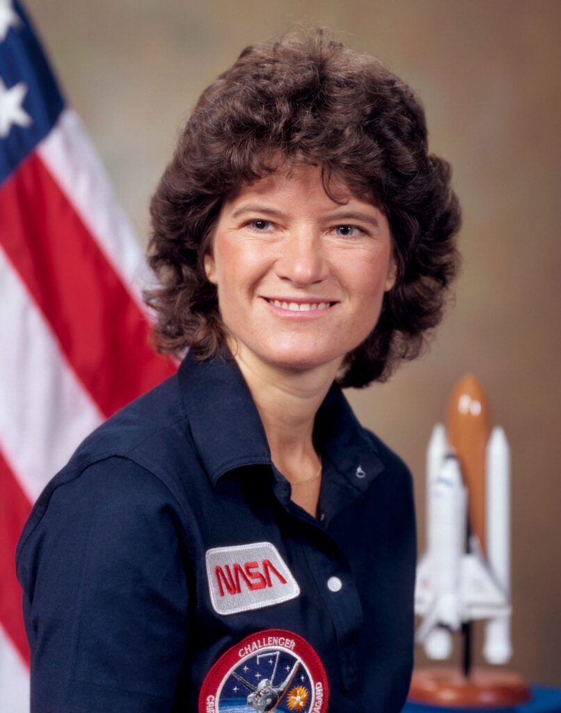 Foto oficial de perfil de Sally Ride, com um sorriso tranquilo. Ao fundo, é possível ver a bandeira dos EUA e uma réplica em miniatura do ônibus espacial, simbolizando sua importante contribuição para as missões espaciais e o legado histórico que deixou como a primeira mulher americana no espaço por esse feito ela é um dos varios astronautas famosos da nasa.