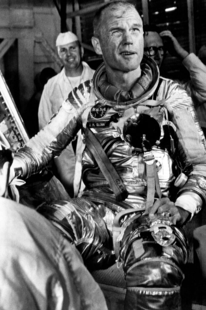 Fotografia em preto e branco de um astronauta famoso da NASA, sorrindo enquanto usa o traje espacial da época. O ambiente parece ser uma sala de comando, com equipamentos ao fundo, capturando um momento de descontração e preparação para a missão. A imagem evoca a era pioneira da exploração espacial.