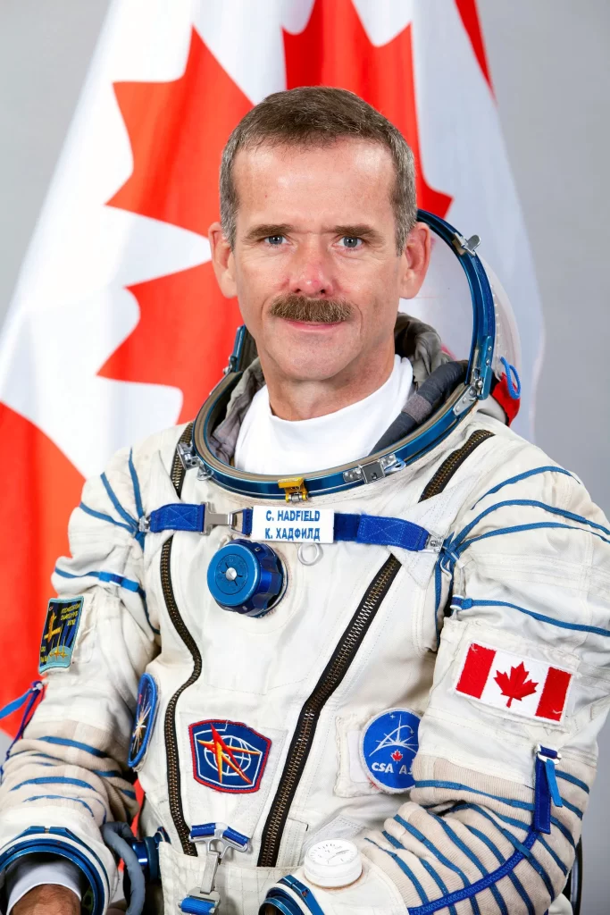 Foto oficial de Chris Hadfield, um dos Astronautas Famosos da NASA, vestindo um traje espacial branco, sem capacete. O logo do Canadá é visível em seu traje, e ao fundo, a parede branca exibe a bandeira do Canadá, refletindo sua origem e a importância de suas contribuições para as missões espaciais da NASA.