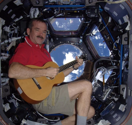 Chris Hadfield, um dos Astronautas Famosos da NASA, usando short creme, camiseta vermelha e meias, tocando violão dentro da Estação Espacial Internacional (ISS). Ao fundo, a janela da estação exibe a atmosfera da Terra, capturando um momento de descontração e humanidade no espaço.