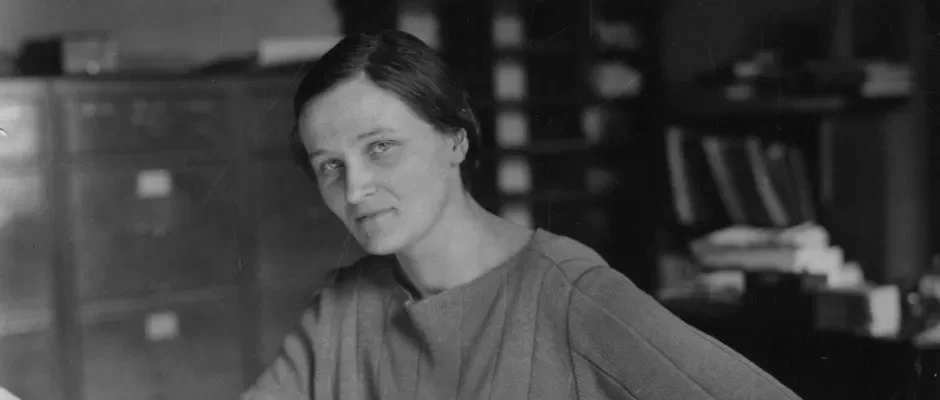 Cecilia Payne-Gaposchkin sentada em uma cadeira, apoiada em uma mesa de escritório, usando roupas da época. A cena é ambientada em uma sala de aparência clássica, com móveis antigos, refletindo o contexto histórico de sua época.