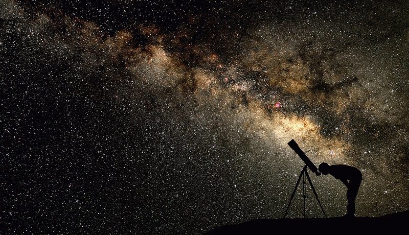 Pessoa observando o céu estrelado com um telescópio, em uma noite clara. A imagem ilustra como utilizar um telescópio para explorar estrelas e outros corpos celestes no céu.