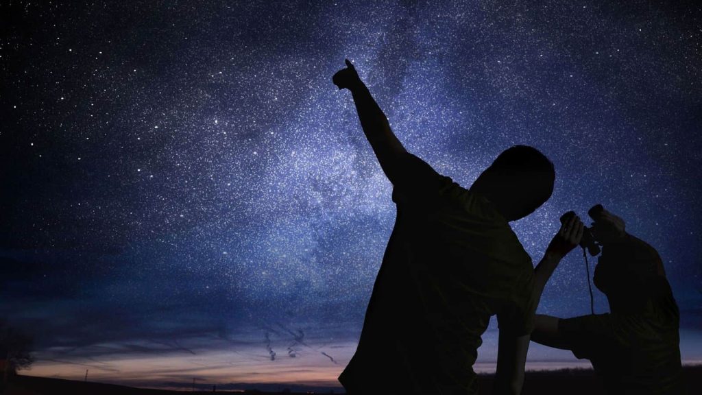 Duas pessoas observando as estrelas com binóculos astronômicos em uma noite de céu limpo. A cena representa uma forma prática de como observar o céu com equipamentos ou a olho nu permitindo explorar constelações e planetas visíveis a olho nu.