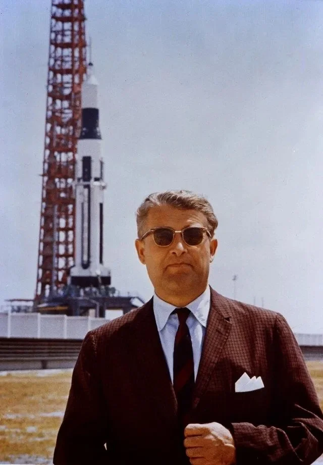 Dr. Wernher von Braun, arquiteto-chefe do foguete, vestindo um terno e óculos de proteção solar escuros, observa atentamente o lançamento do Apollo Saturn V ao fundo em um dia ensolarado