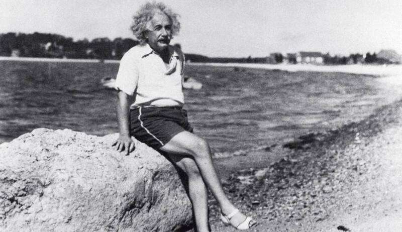 Albert Einstein em um momento de descanso no Rio de Janeiro, Brasil. Ele está sentado de pernas cruzadas em uma grande pedra, vestindo uma camiseta branca e shorts, com uma expressão serena no rosto. O físico parece em paz, olhando para o horizonte com um olhar calmo e contemplativo. Ao fundo, é possível ver as montanhas e a vegetação típica do Rio de Janeiro, sugerindo um ambiente tranquilo e natural. A cena transmite um senso de serenidade e conexão com a natureza.