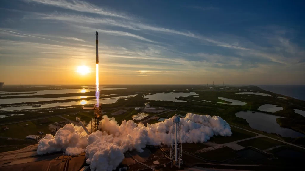 Foguete Falcon 9 sendo lançado de uma base espacial, com a propulsão gerando uma grande coluna de fumaça e fogo. A cena captura o momento inicial do lançamento, destacando a potência do foguete em direção ao céu. A palavra-chave "Lançamento espacial" é representada pela ação de ascensão do foguete.