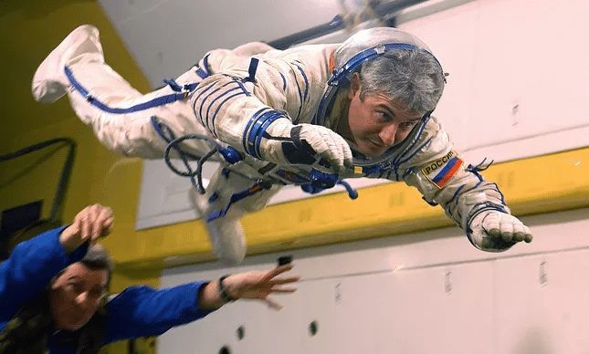 Marcos Pontes, astronauta brasileiro, sorri em sua foto oficial da NASA. Ele está usando um traje espacial branco, com detalhes em azul e vermelho, e não está usando o capacete. Seu traje possui a bandeira do Brasil no ombro esquerdo. Ao fundo, a bandeira do Brasil está visível, destacando a sua nacionalidade. A imagem transmite um ar de orgulho e conquista.