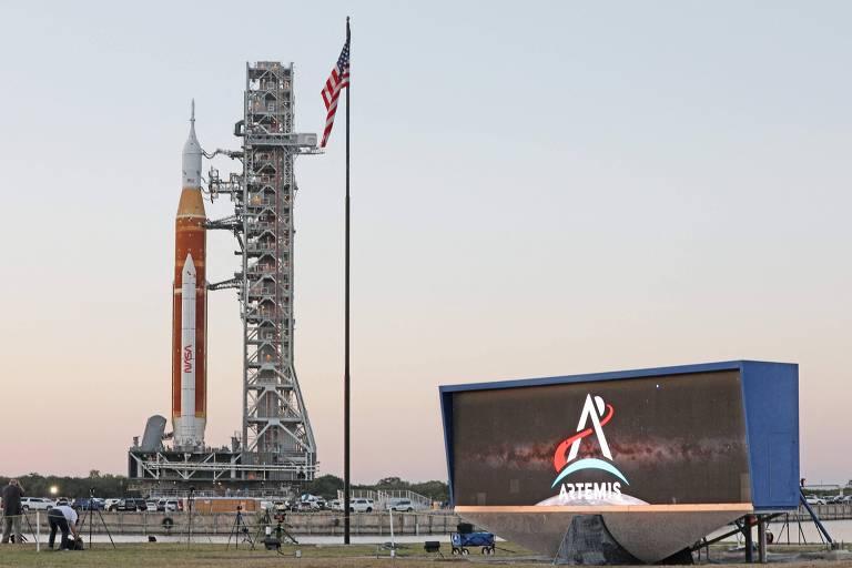 Vista distante do foguete SLS (Space Launch System) da NASA, imponente na plataforma de lançamento. O tanque laranja é destacado, com a bandeira dos EUA hasteada no mastro ao lado. Um banner digital da NASA com o logo do programa Artemis também é visível. A foto foi tirada ao entardecer, com o céu criando uma iluminação dramática que realça o tamanho e a grandiosidade do foguete.