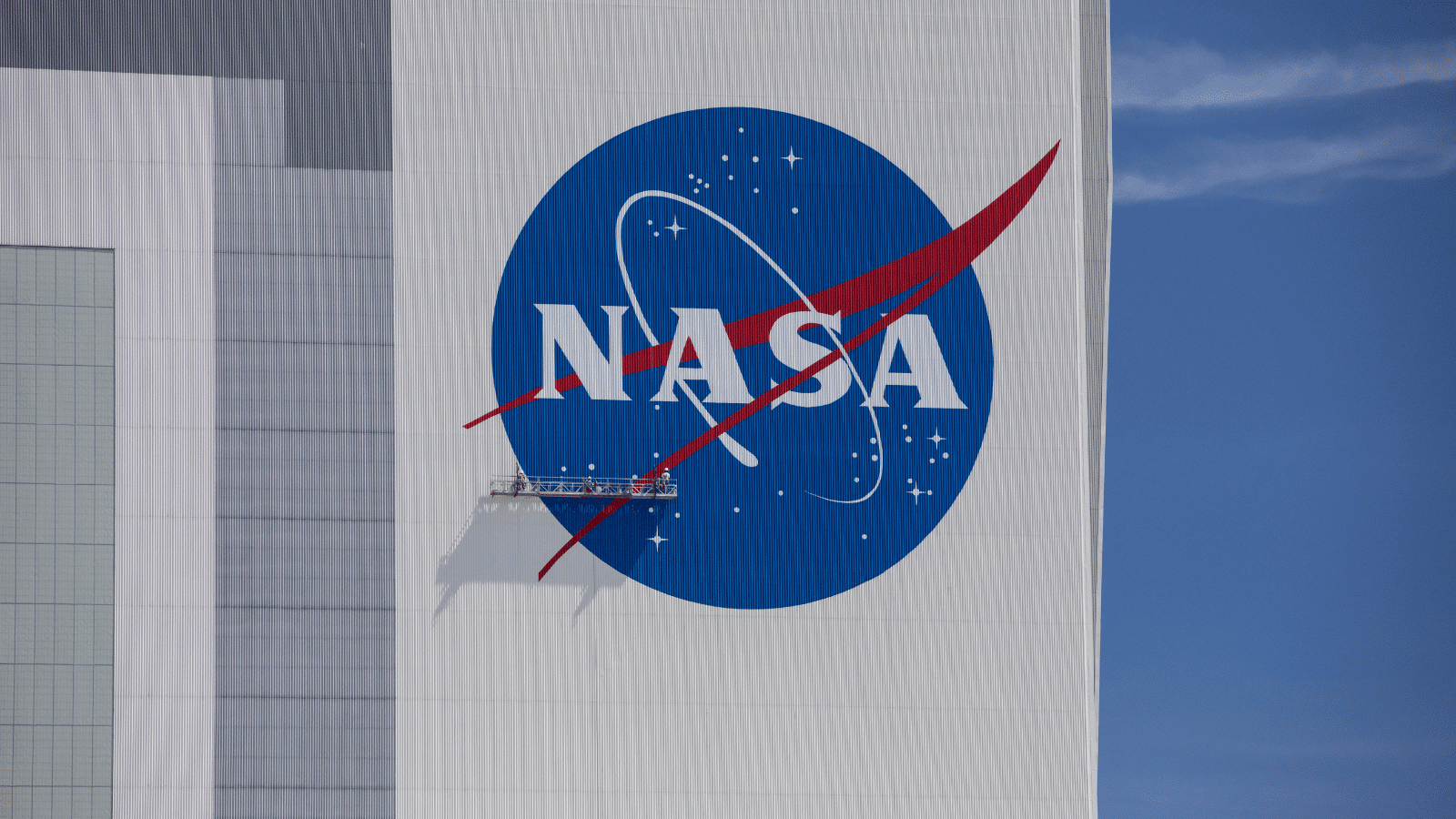 Imagem do logo da NASA na fachada do Kennedy Space Center, com trabalhadores realizando manutenção na área. O logo, de grande escala, ocupa grande parte da fachada, e os trabalhadores, em tamanho reduzido devido à magnitude do logo, parecem minúsculos em comparação com a imponente estrutura. A cena transmite a grandiosidade do local e a dedicação dos profissionais envolvidos na conservação do símbolo.