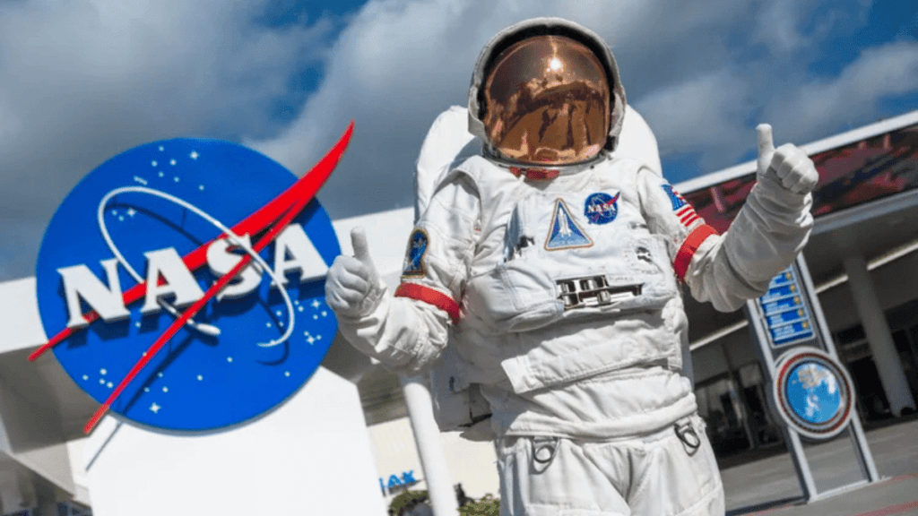 ecepção do Kennedy Space Center, com o logo grande da NASA ao fundo. Em primeiro plano, um astronauta usando traje espacial faz o sinal de 'joia' com as mãos, convidando os visitantes a conhecerem mais sobre o que é a NASA, onde fica e como funciona
