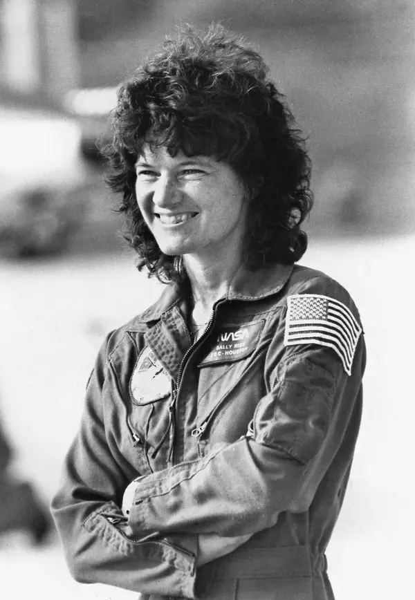 Astronautas famosos da NASA Fotografia de Sally Ride sorrindo, com os braços cruzados e olhando para a frente. Ela usa o traje espacial da NASA, com a bandeira dos EUA visível em seu ombro, transmitindo confiança e orgulho por sua histórica conquista como a primeira mulher americana no espaço.