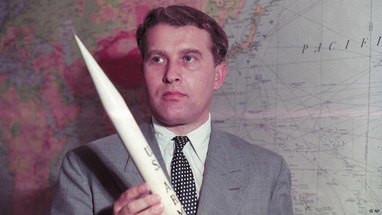 "Wernher von Braun jovem, de olhos claros, vestindo um terno cinza e camiseta branca, segura uma miniatura de foguete com a sigla 'US Army'. Ao fundo, um mapa-múndi destaca sua influência global na engenharia de foguetes.