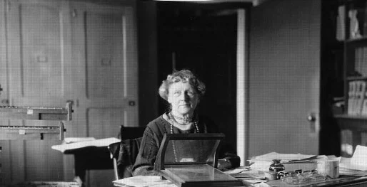 Annie Jump Cannon, astrônoma pioneira, sentada em sua mesa de trabalho, com livros e observações astronômicas ao seu redor. Ela está analisando estrelas e classificando-as, com um olhar focado e concentrado. Ao fundo, um quadro de constelações e um telescópio, simbolizando suas contribuições para a astronomia, especialmente sua criação do sistema de classificação espectral das estrelas.