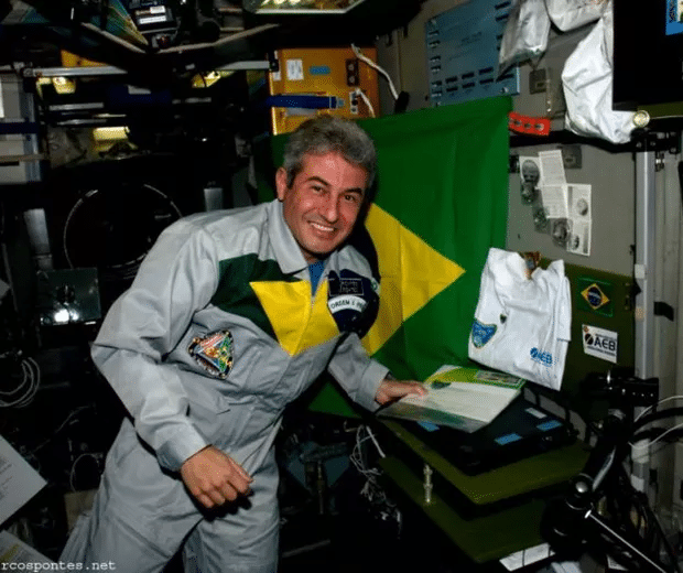 Foto de Marcos Pontes, astronauta brasileiro, dentro da Estação Espacial Internacional (ISS). Ele usa um traje espacial cinza com a bandeira do Brasil como uma faixa no peito. Próximo a ele, estão alguns equipamentos da ISS, e ele está próximo a uma mesa onde há um notebook preto com um adesivo da AEB (Agência Espacial Brasileira).