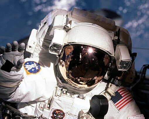 Patrick G. Forrester, especialista da missão STS-105, cumprimenta um membro da tripulação dentro da cabine do ônibus espacial Discovery durante uma das sessões de atividade extraveicular (EVA), em 2001. Ao fundo, é possível ver a curvatura da Terra, destacando a perspectiva única do espaço.