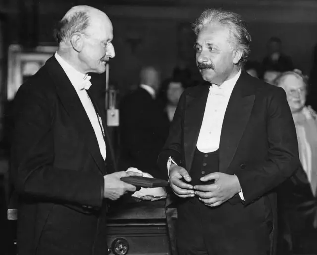 Max Planck e Albert Einstein de pé, frente a frente, ambos usando trajes formais. Eles estão em uma conversa animada sobre física, com expressões sérias e envolvidas, e gesticulando com as mãos enquanto discutem conceitos científicos profundos. A cena transmite um momento histórico de troca intelectual entre os dois grandes físicos