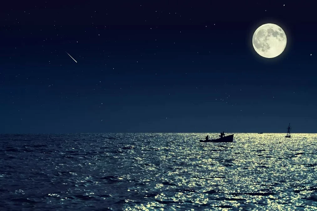 Curiosidades sobre a Lua: Em uma cena noturna deslumbrante, a Lua cheia brilha intensamente no céu estrelado, refletindo sua luz prateada sobre as águas tranquilas do mar. No horizonte, a silhueta de um barco de pesca adiciona profundidade à paisagem, enquanto uma estrela cadente risca o céu, criando um momento de pura beleza e mistério.