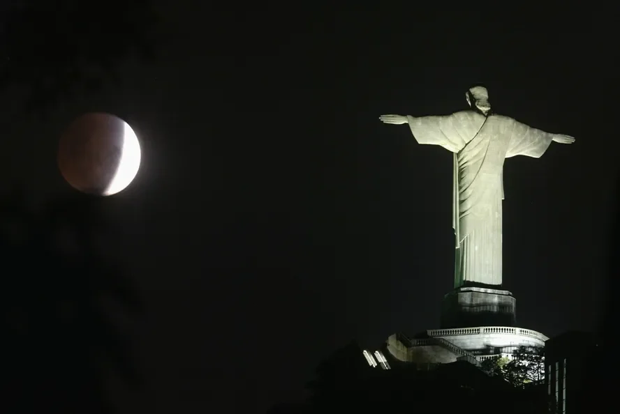 eclipse lunar, com a Lua visível ao fundo, brilhando intensamente no céu noturno. O Cristo Redentor do Rio de Janeiro aparece desfocado na frente, com sua silhueta suavemente iluminada pela luz da Lua, criando um contraste suave entre o monumento e o céu.