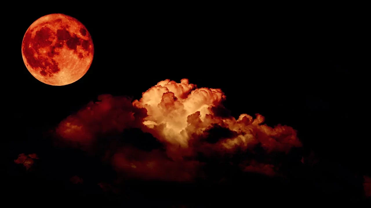 eclipse lunar total, conhecido como Lua de Sangue. A Lua aparece à direita da imagem, com uma tonalidade avermelhada intensa. Do lado esquerdo, grandes nuvens ocupam parte do céu, refletindo algumas tonalidades vermelhas devido à luz dispersa na atmosfera.
