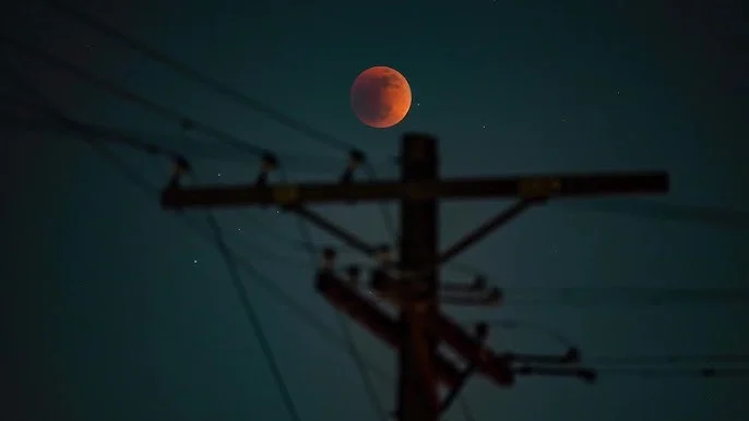Imagem do eclipse lunar total, conhecido como Lua de Sangue, vista de uma rua. A Lua avermelhada aparece atrás de postes e fios elétricos, criando um contraste interessante entre o fenômeno celestial e o cenário urbano.