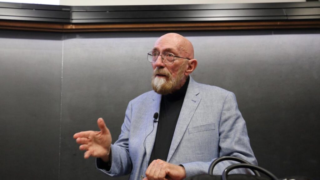 Kip Thorne em uma pose de perfil, com olhar concentrado e expressão séria. Ele veste um terno escuro, e a iluminação destaca os traços marcantes de seu rosto, refletindo sua profunda dedicação à física teórica.