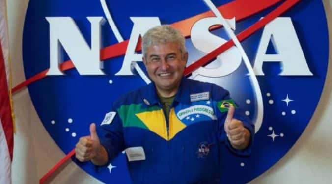 Marcos Pontes, astronauta brasileiro, posa em frente ao logo da NASA. Ele está vestindo um traje espacial azul, com a bandeira do Brasil no peito, e faz o sinal de 'joia' com ambas as mãos.