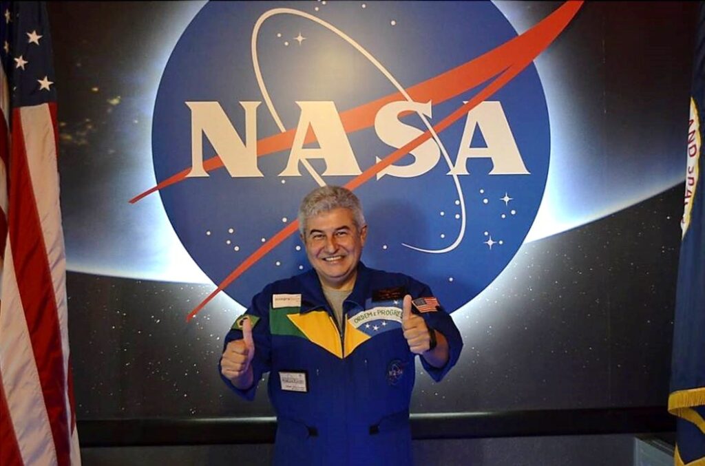 Marcos Pontes, astronauta brasileiro, posa em frente ao logo da NASA. Ele está vestindo um traje espacial azul, com a bandeira do Brasil no peito, e faz o sinal de 'joia' com uma mão