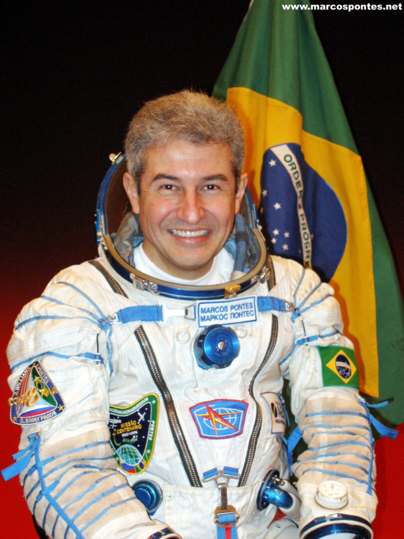 Marcos Pontes, astronauta brasileiro, sorri em sua foto oficial da NASA. Ele está usando um traje espacial branco, com detalhes em azul e vermelho, e não está usando o capacete. Seu traje possui a bandeira do Brasil no ombro esquerdo. Ao fundo, a bandeira do Brasil está visível, destacando a sua nacionalidade. A imagem transmite um ar de orgulho e conquista.