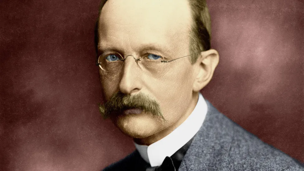 Max Planck, com olhos claros, bigode e óculos, exalando um olhar sério. Ele está vestido com um terno cinza e gravata preta, capturando sua postura de autoridade e intelectualidade.