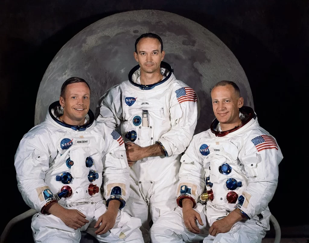 A tripulação da Apollo 11 — Neil Armstrong, Michael Collins e Edwin 'Buzz' Aldrin — posando em trajes espaciais sem capacete, com uma imagem da Lua ao fundo. A missão Apollo 11 foi a primeira a levar humanos à superfície lunar, marcando um momento histórico na exploração espacial."